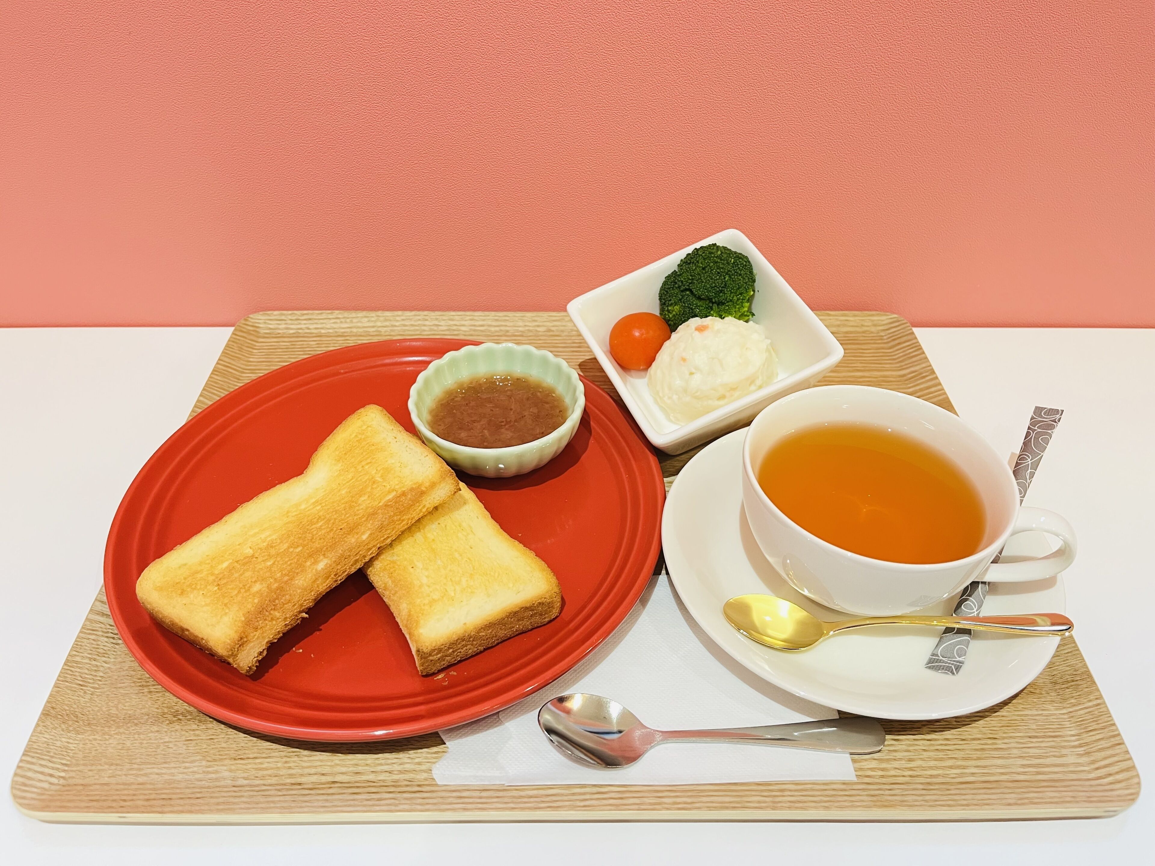 A meal with toast, soup, and vegetables on a red plate and a cup of tea on a white saucer.