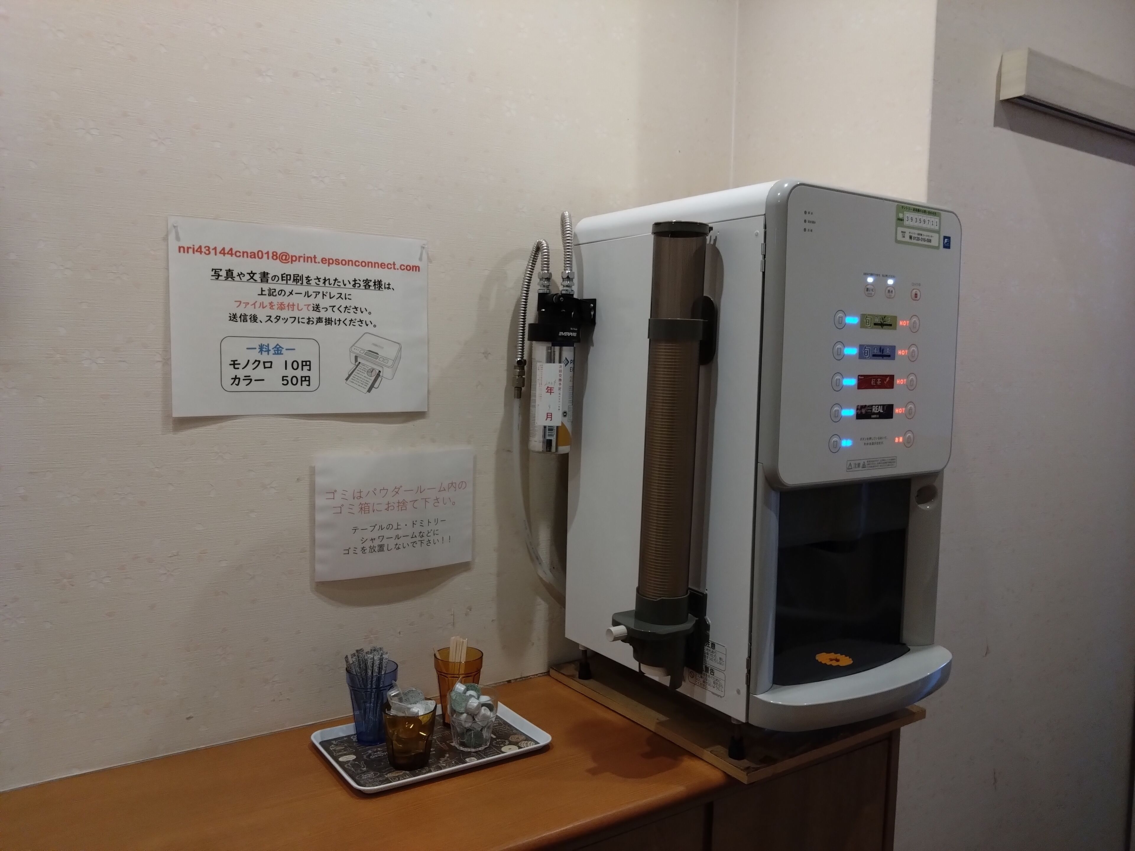 A water dispenser with a control panel, a receipt holder, and a tray with cups and a thermos.