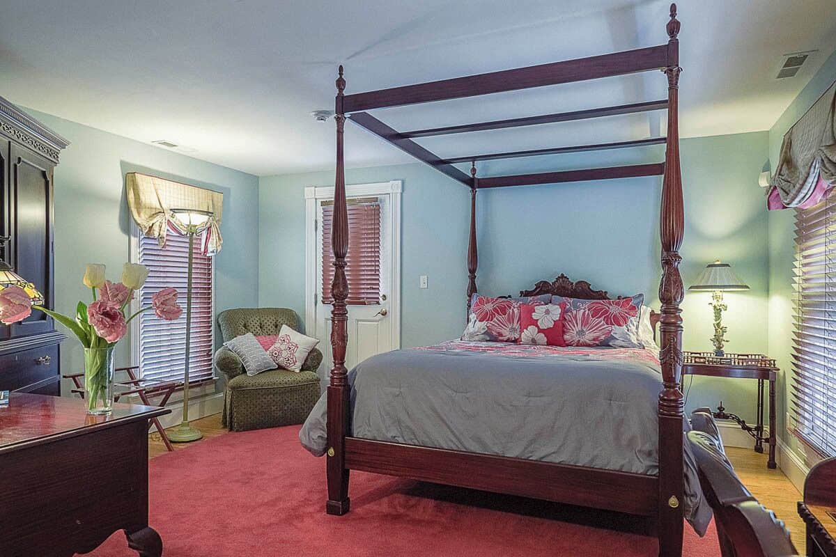 A four-poster bed in a room with a red carpet, a desk, a chair, and a small table with a lamp.