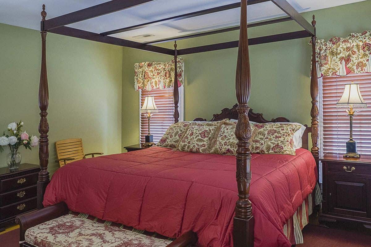A four-poster bed with a red bedspread, floral-patterned pillows, and a matching footboard. There are two nightstands with lamps and a dresser with drawers.