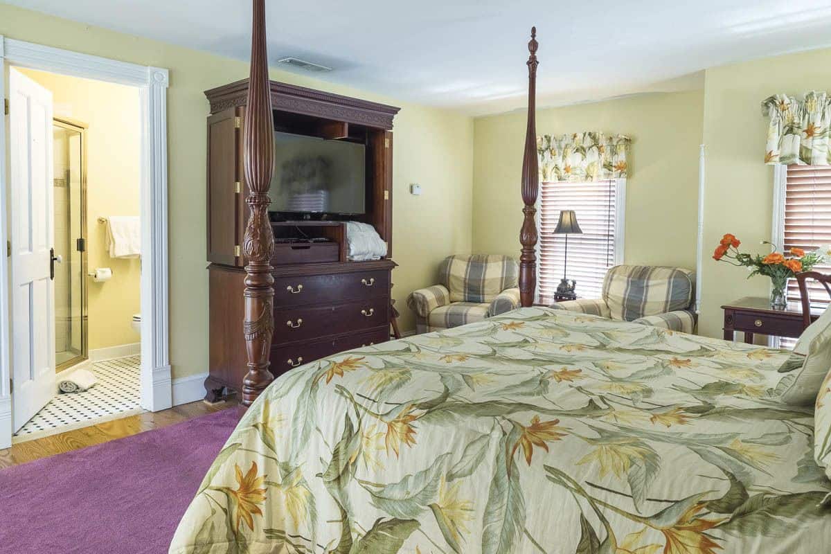 A bedroom with a four-poster bed, a wooden dresser, a television, and a window with blinds.