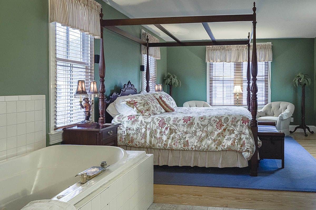 A four-poster bed with a floral bedspread, a large bathtub, and a chair in a room with green walls and wooden floors.