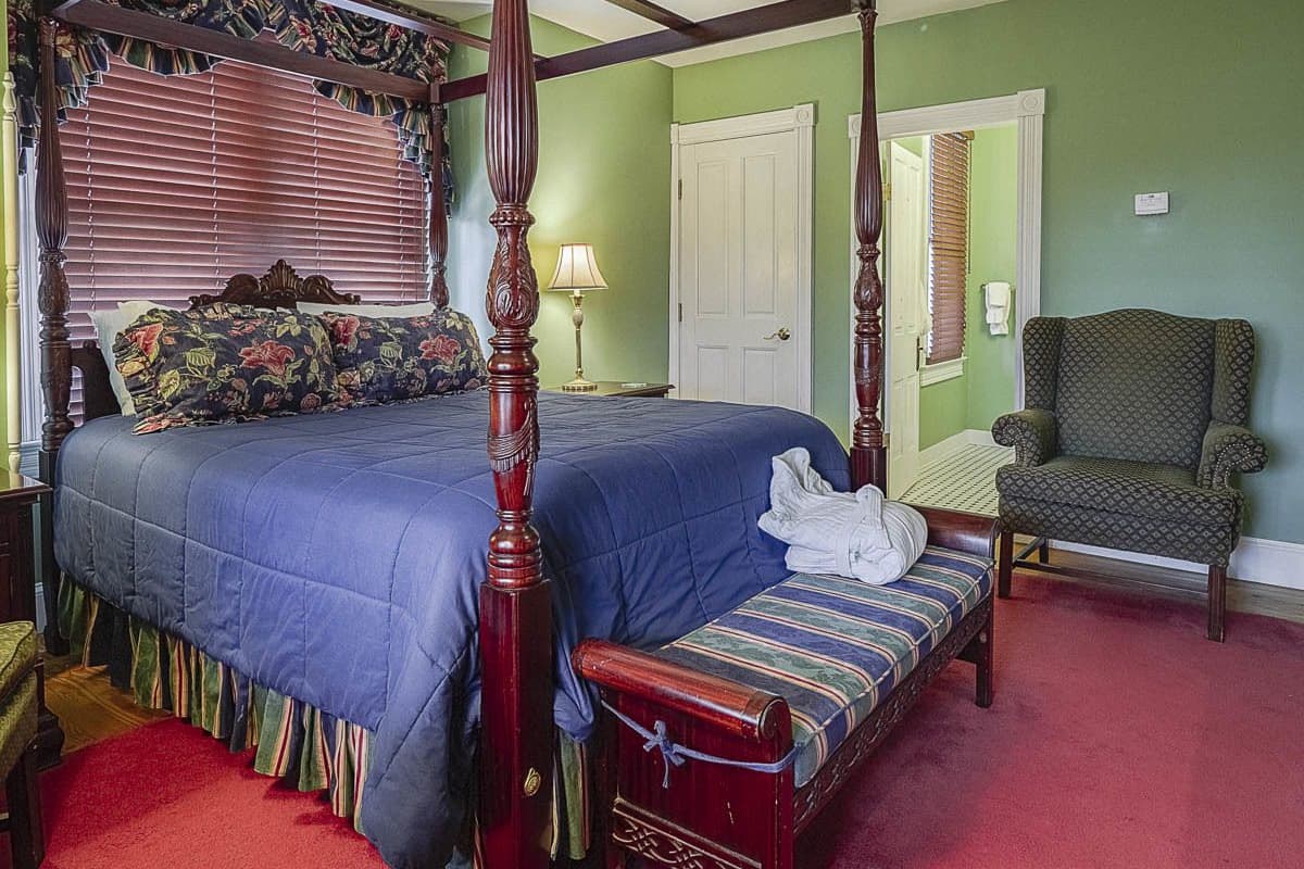A four-poster bed with a blue bedspread and floral pillows, a matching bench with a towel, a green armchair, and a white door.