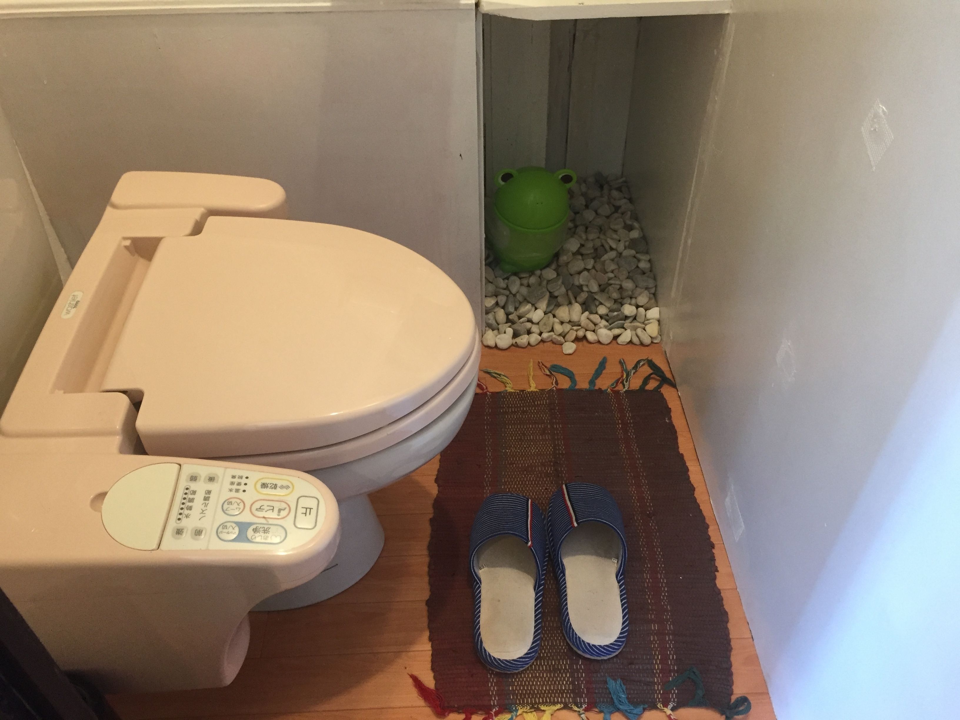 A toilet with a control panel, a green bucket, and a pair of slippers on a woven mat.