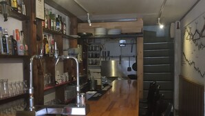 Eine Bar mit Holztresen, Hockstühlen, Regalen mit Flaschen und Küchenausstattung im Hintergrund.