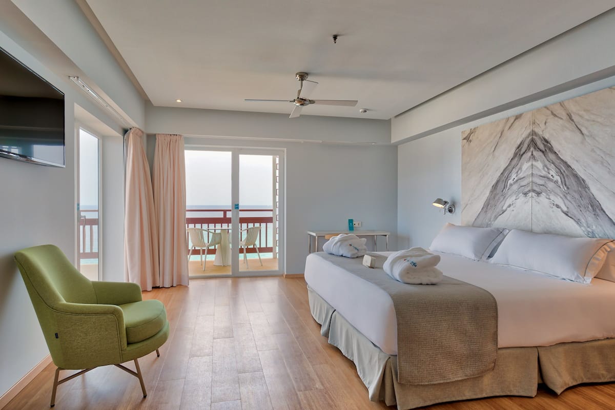 A hotel room with a bed, a green chair, a desk, a large marble wall, and a balcony with a view.