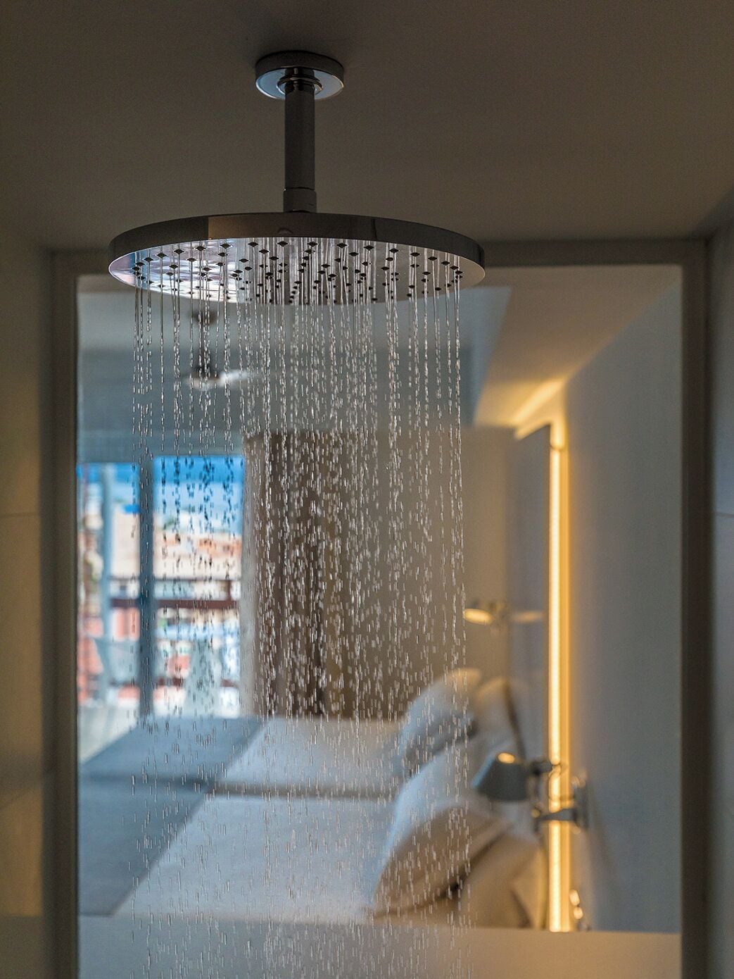 A modern bathroom with a rain showerhead, a visible window, and a bed with a nightstand.