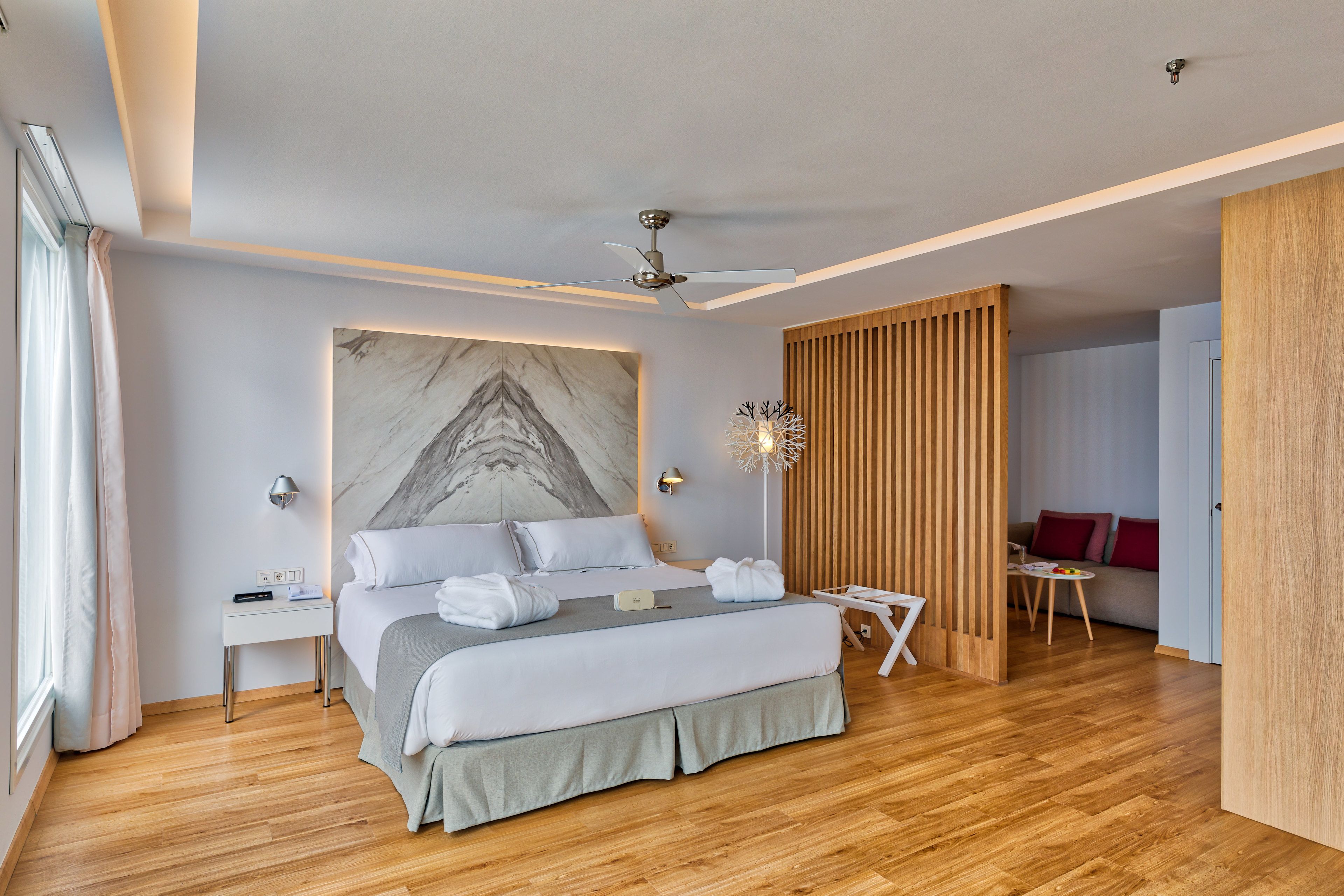 A modern bedroom with a large bed, a ceiling fan, a marble artwork, and a wooden partition.
