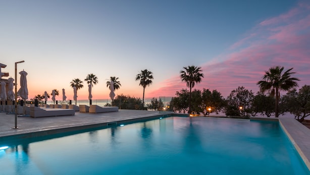 Zona de piscina con sillones de descanso y sombrillas, palmeras y vista al atardecer.