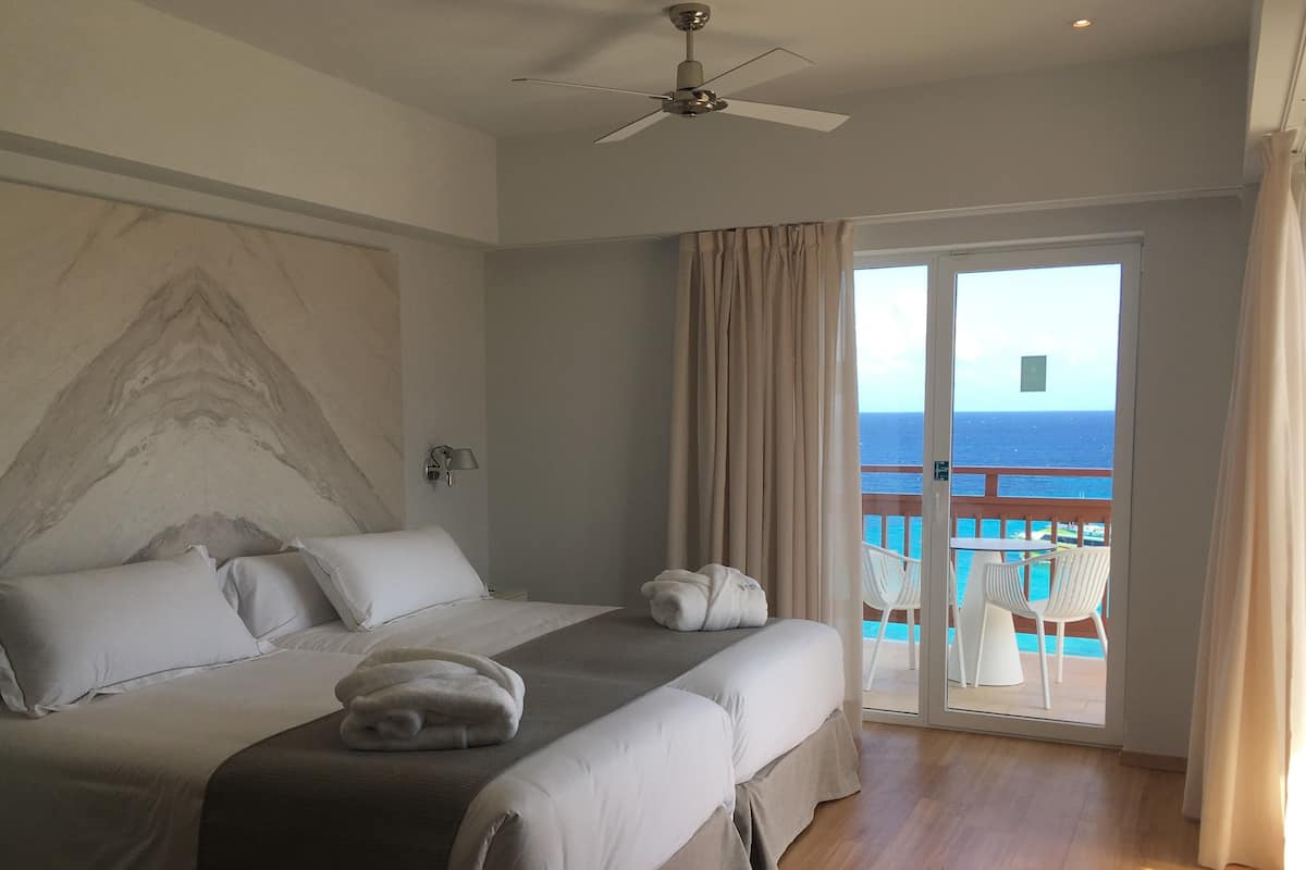 A hotel room with a bed, a ceiling fan, a view of the ocean, and a balcony with white chairs.