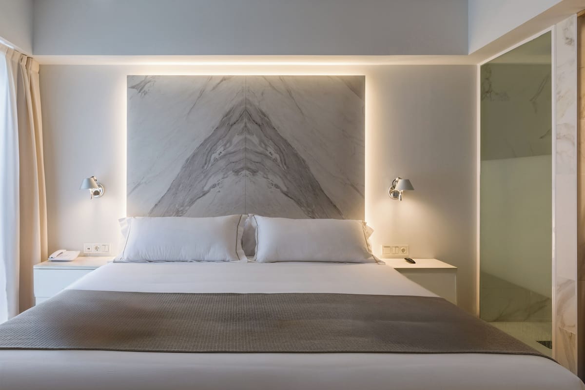 Une chambre d’hôtel moderne dotée d’un grand lit, de deux tables de chevet, d’une tête de lit en marbre et offrant une vue sur la ville.