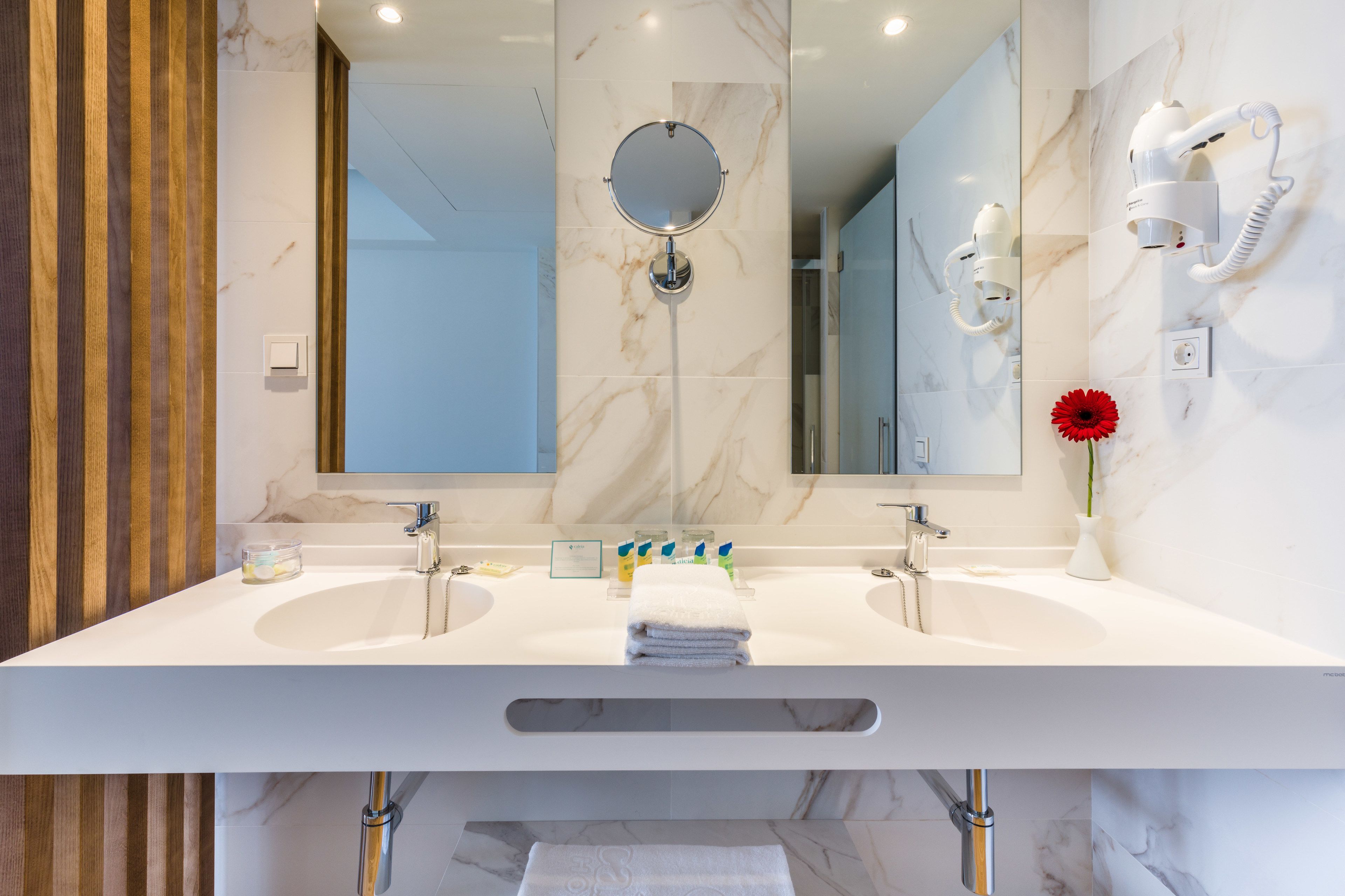 A modern bathroom with two sinks, a large mirror, a hairdryer, and a vase with a red flower.