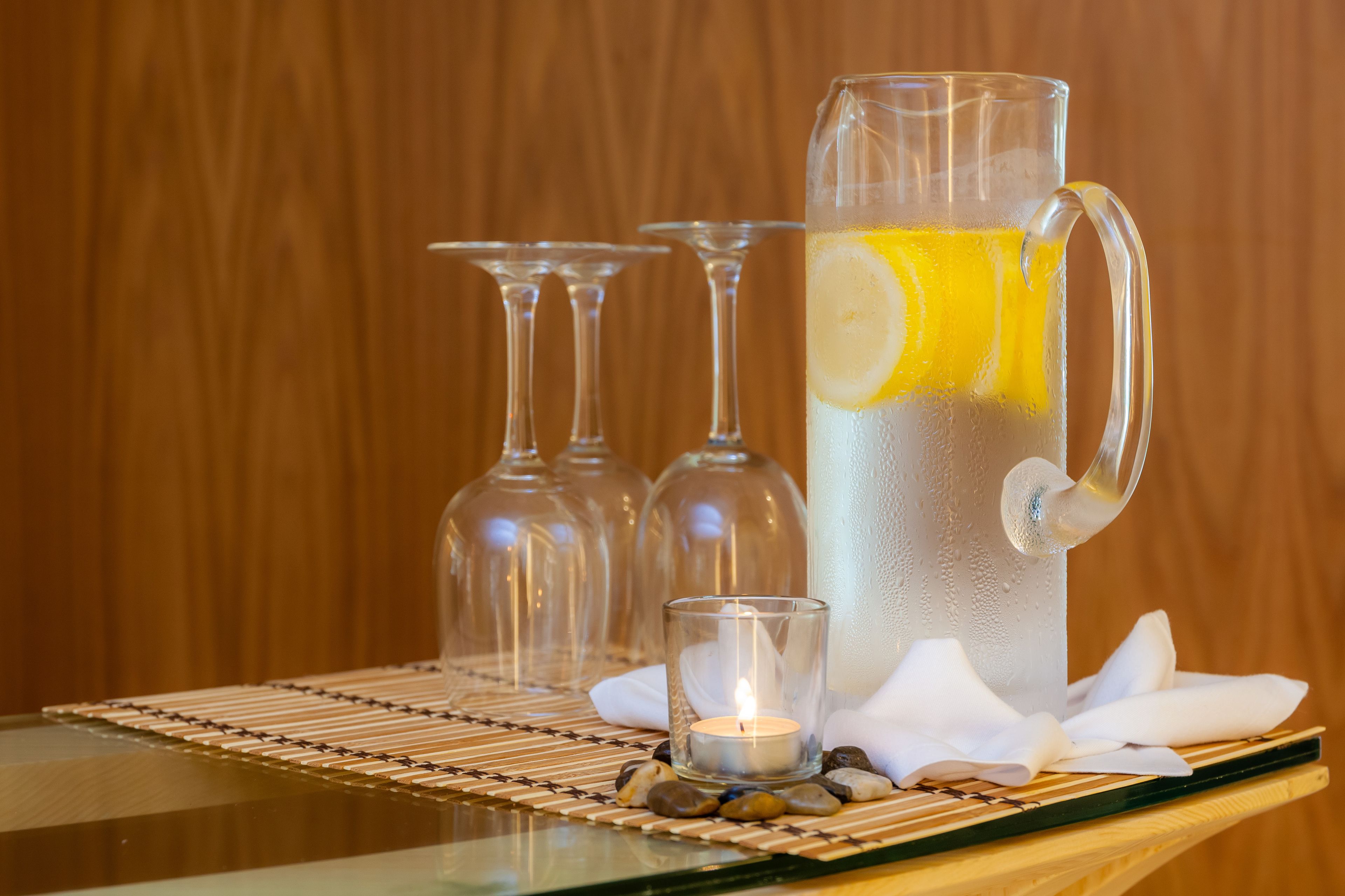Un jarro de vidrio transparente con una rodaja de limón, una vela encendida y dos copas de vino sobre una mesa de madera.