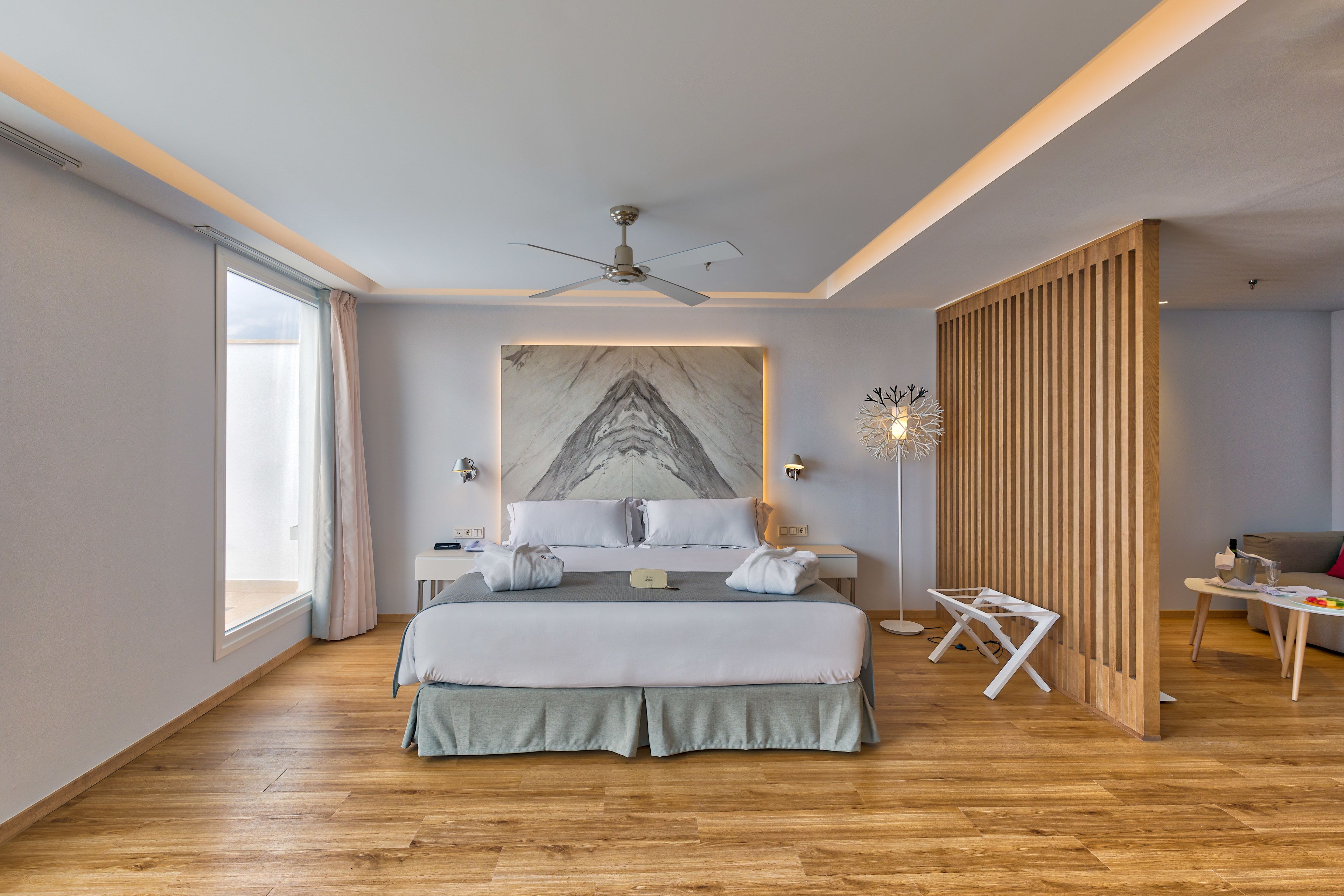 A modern bedroom with a large bed, a ceiling fan, a marble artwork, and a wooden partition.