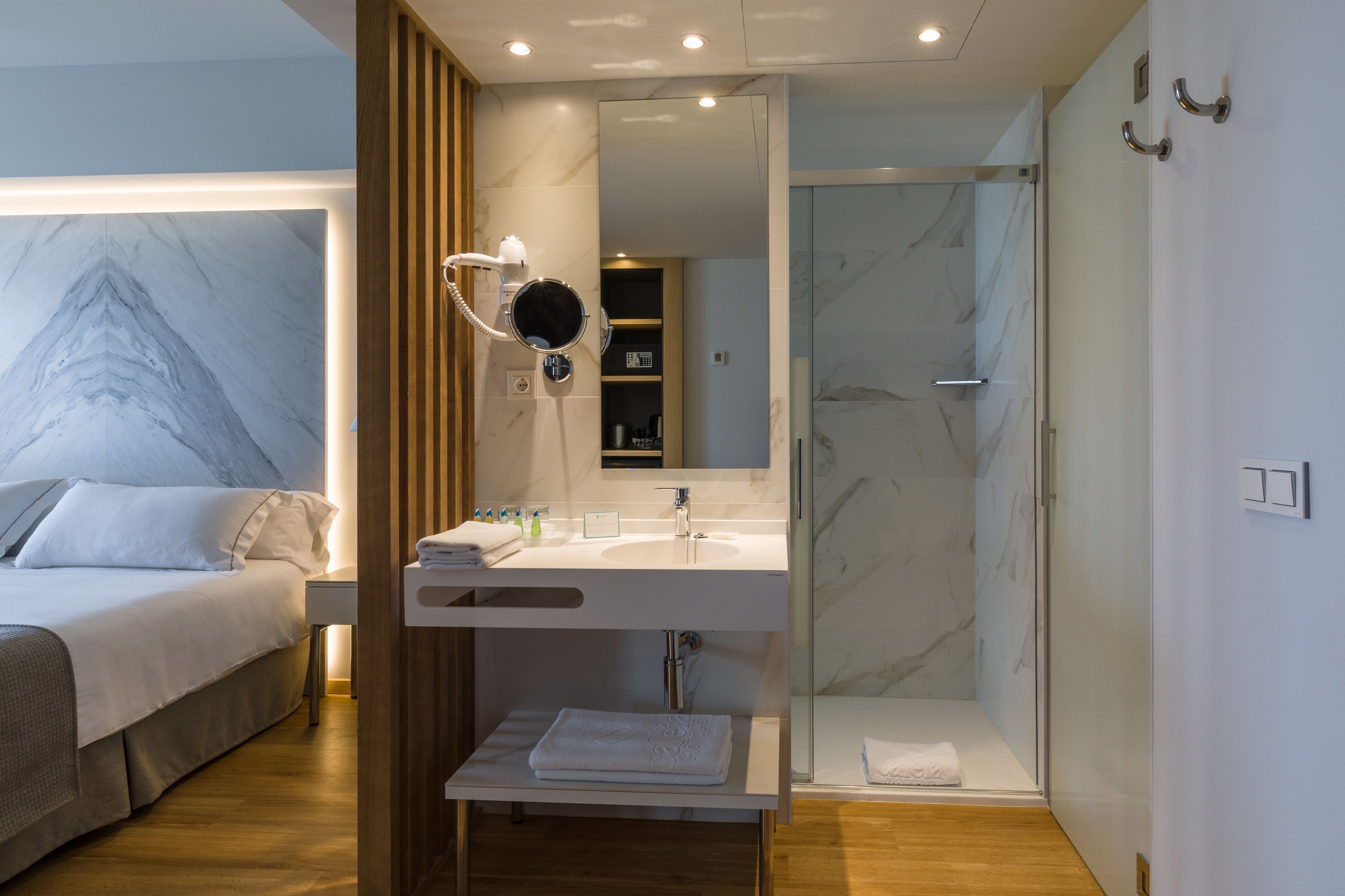 A modern hotel room with a bed, a bathroom with a shower, and a vanity with a mirror.