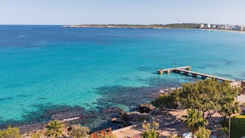 Vista costera con un muelle que se adentra en aguas turquesas, un litoral rocoso y un edificio con terraza.