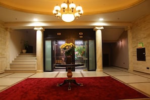 A lobby with a red carpet, a chandelier, a staircase, and a reception desk with a vase of flowers.