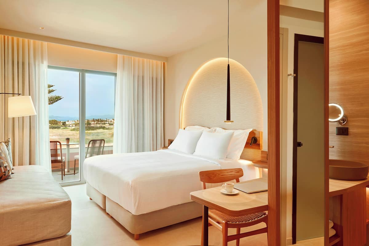 Habitación de hotel con una cama grande, un escritorio con silla, una lámpara y vistas al exterior.