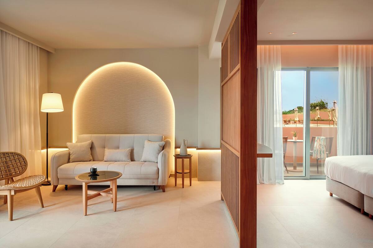 Habitación de hotel moderna con sofá, una mesita, lámpara y cama. Cuenta con un gran ventanal con cortinas y vistas al exterior.