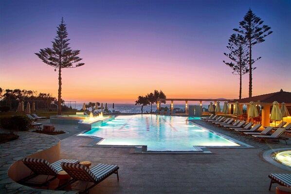Un espace piscine doté de chaises longues et de parasols, offrant une vue sur l’océan au coucher du soleil.