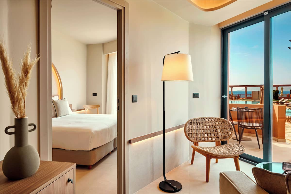 Una habitación de hotel moderna con cama, una zona de estar con una silla y una mesa, una lámpara de pie y vista al mar a través de amplias ventanas.