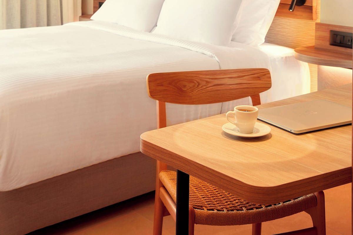Una habitación de hotel con una cama, una mesa de madera con una computadora portátil y una taza, y un lámpara colgante.