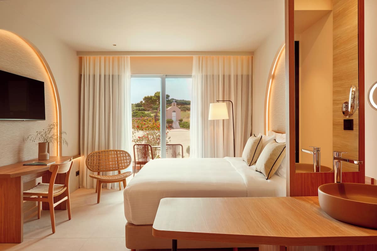 Una habitación de hotel moderna con una cama grande, un escritorio de madera, una silla, una mesita pequeña, un baño con lavamanos y espejo, y vistas al exterior.