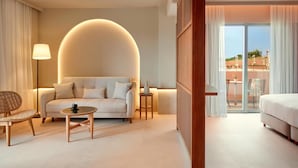A hotel room with a sofa, a chair, a small table, a lamp, and a bed. There is a large window with curtains and a view of the outside. - Zeus Kos Beach (Kos)