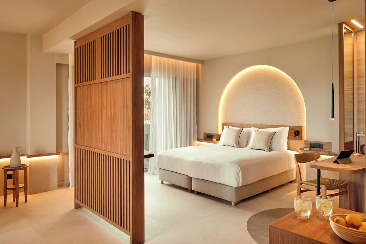 Una habitación de hotel moderna con una cama grande, mesitas de noche, un escritorio con una computadora portátil y un área de comedor con fruta.