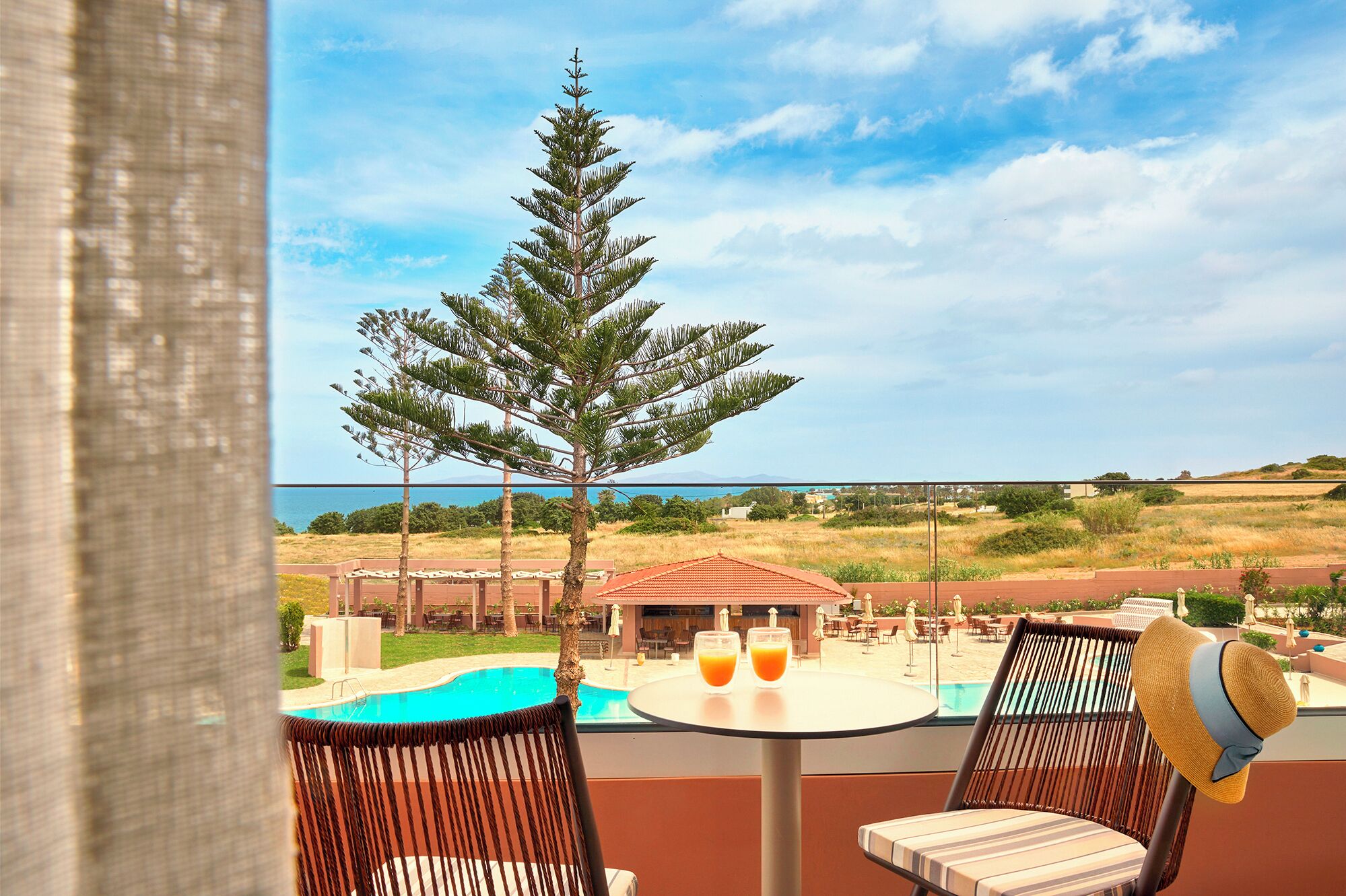 A balcony with a view of a pool, two chairs, a table with drinks, and a large tree.