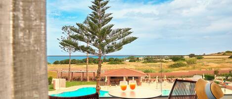 A balcony with a view of a pool, two chairs, a table with drinks, and a large tree.