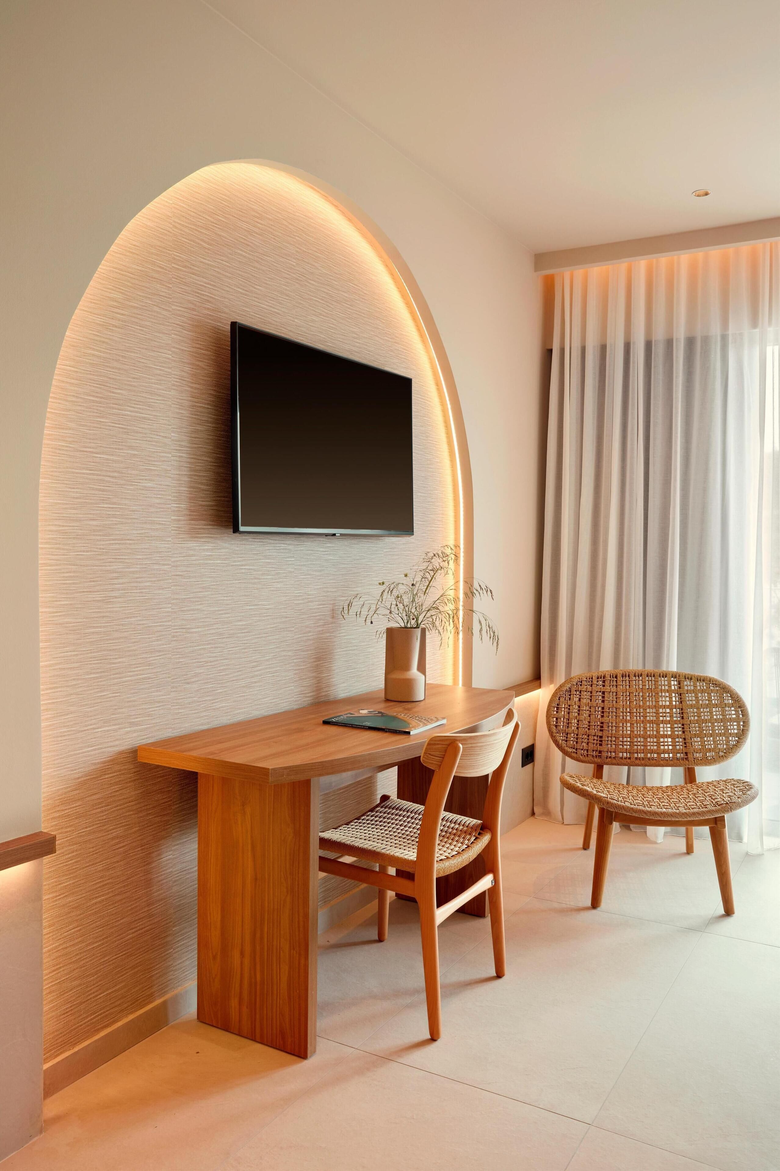 Une chambre d’hôtel moderne avec un bureau en bois, deux fauteuils en osier, un téléviseur à écran plat fixé à un mur texturé et une grande fenêtre ornée de rideaux légers.