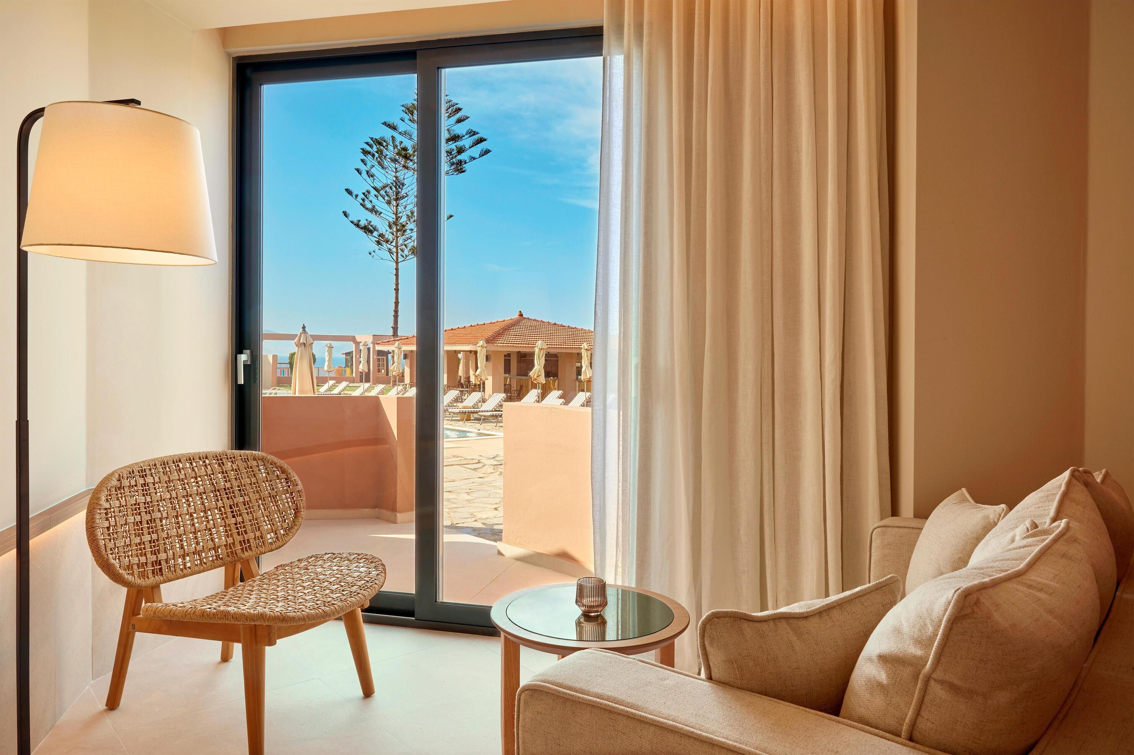 A hotel room with a wicker chair, a sofa, a lamp, and a view of a pool and outdoor seating area.