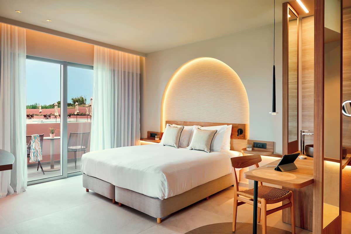Una habitación de hotel moderna con una cama grande, un escritorio con una computadora portátil, una silla y vistas al balcón exterior.