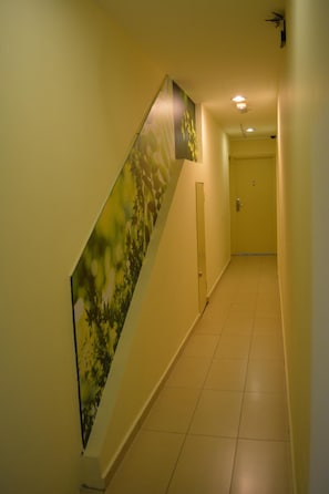 A narrow, well-lit hallway with a door at the end, a wall-mounted light fixture, and a large floral poster on the left wall.