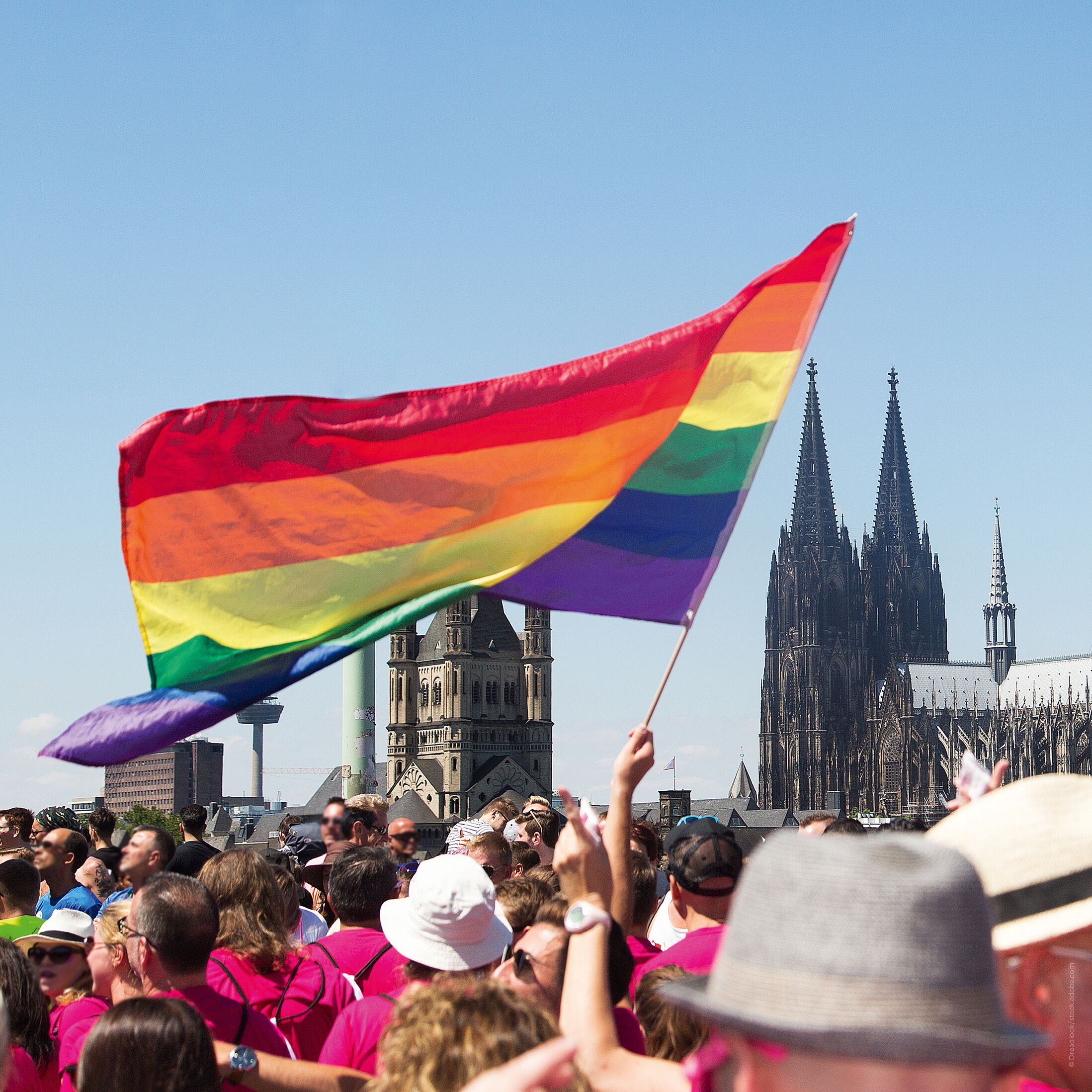 A crowd of people holding a rainbow flag in front of a gothic cathedral.