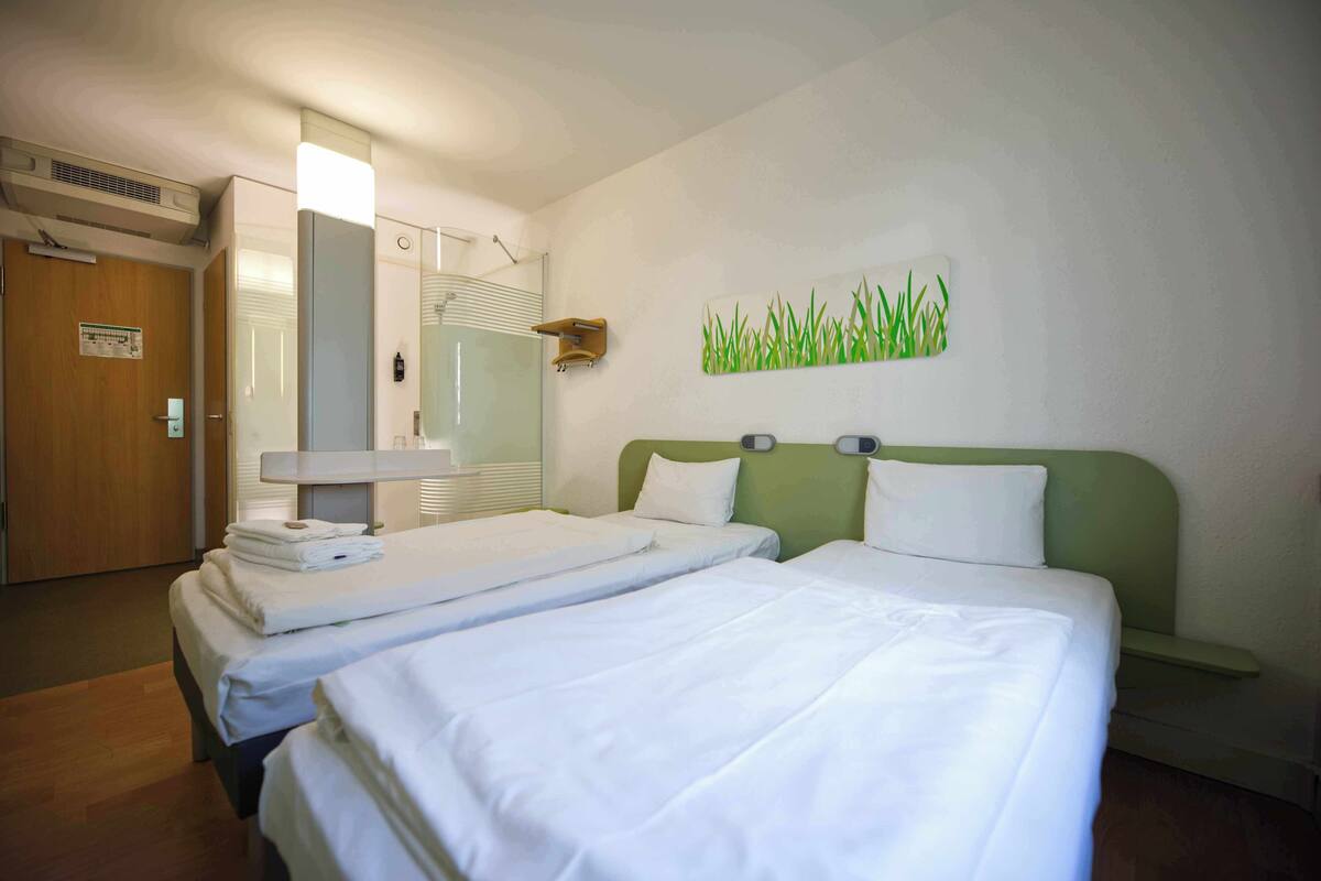 A hotel room with two beds, a wooden door, a mirror, a wall-mounted light, and a green wall art piece.