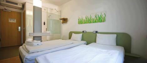 A hotel room with two beds, a wooden door, a mirror, a wall-mounted light, and a green wall art piece.