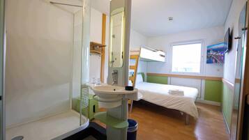 Une chambre d’hôtel compacte, dotée d’une douche, d’un lavabo et d’un lit, et offrant une vue sur le paysage urbain par la fenêtre.