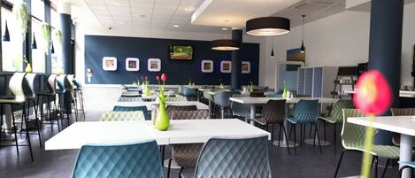 Un restaurante moderno con mesas y sillas blancas, una pared azul oscuro con cuadros enmarcados y plantas colgantes.