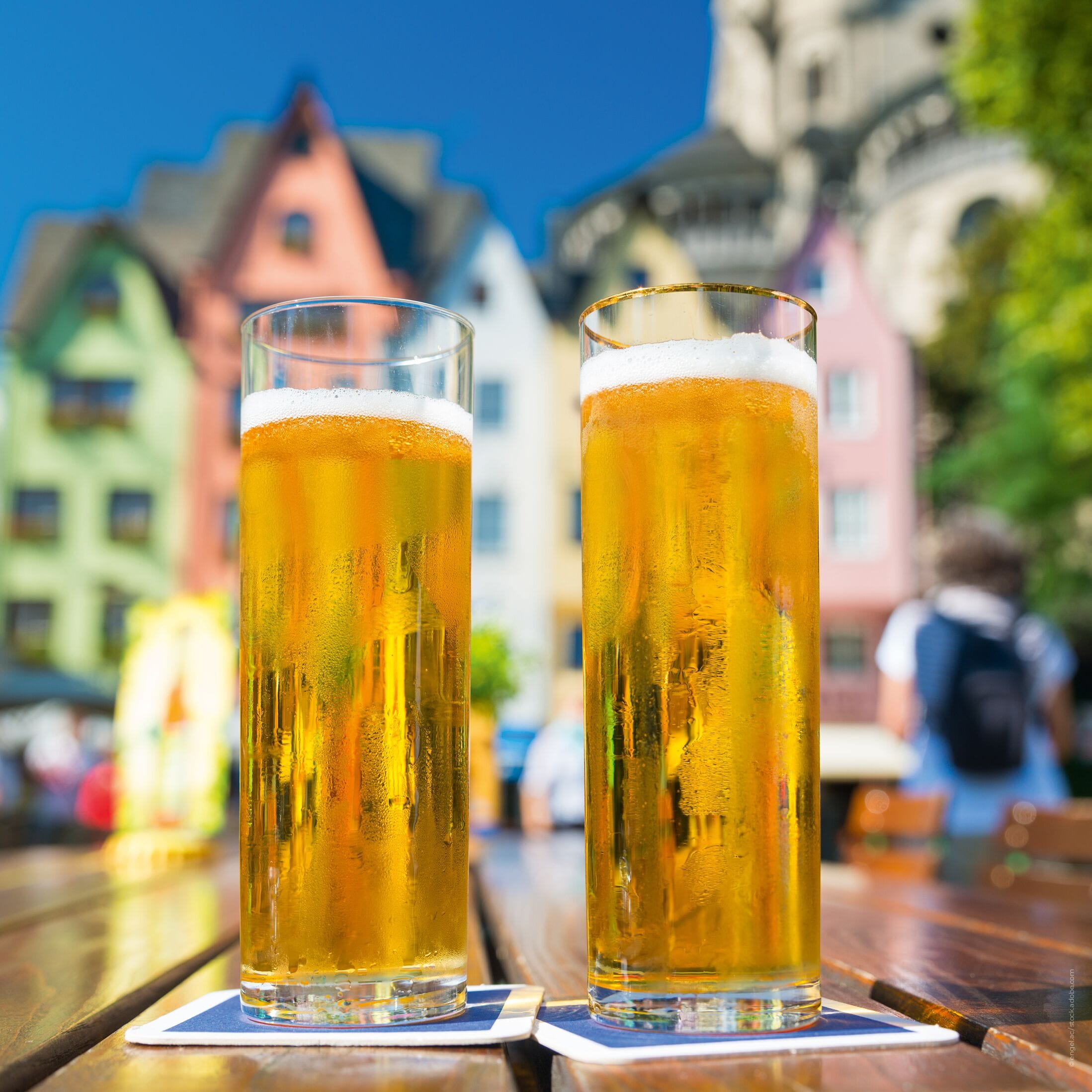 Deux grands verres de bière sur une table en bois, avec en arrière-plan, flou, des bâtiments aux couleurs vives.
