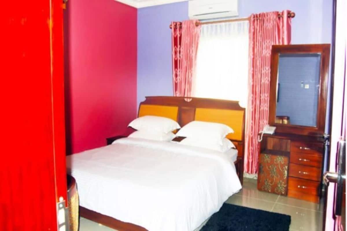 A bedroom with a bed, a wooden dresser, a mirror, and a window with curtains.