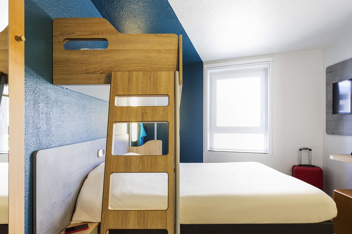 A bunk bed with a wooden ladder, a white mattress, and a red suitcase on the floor.