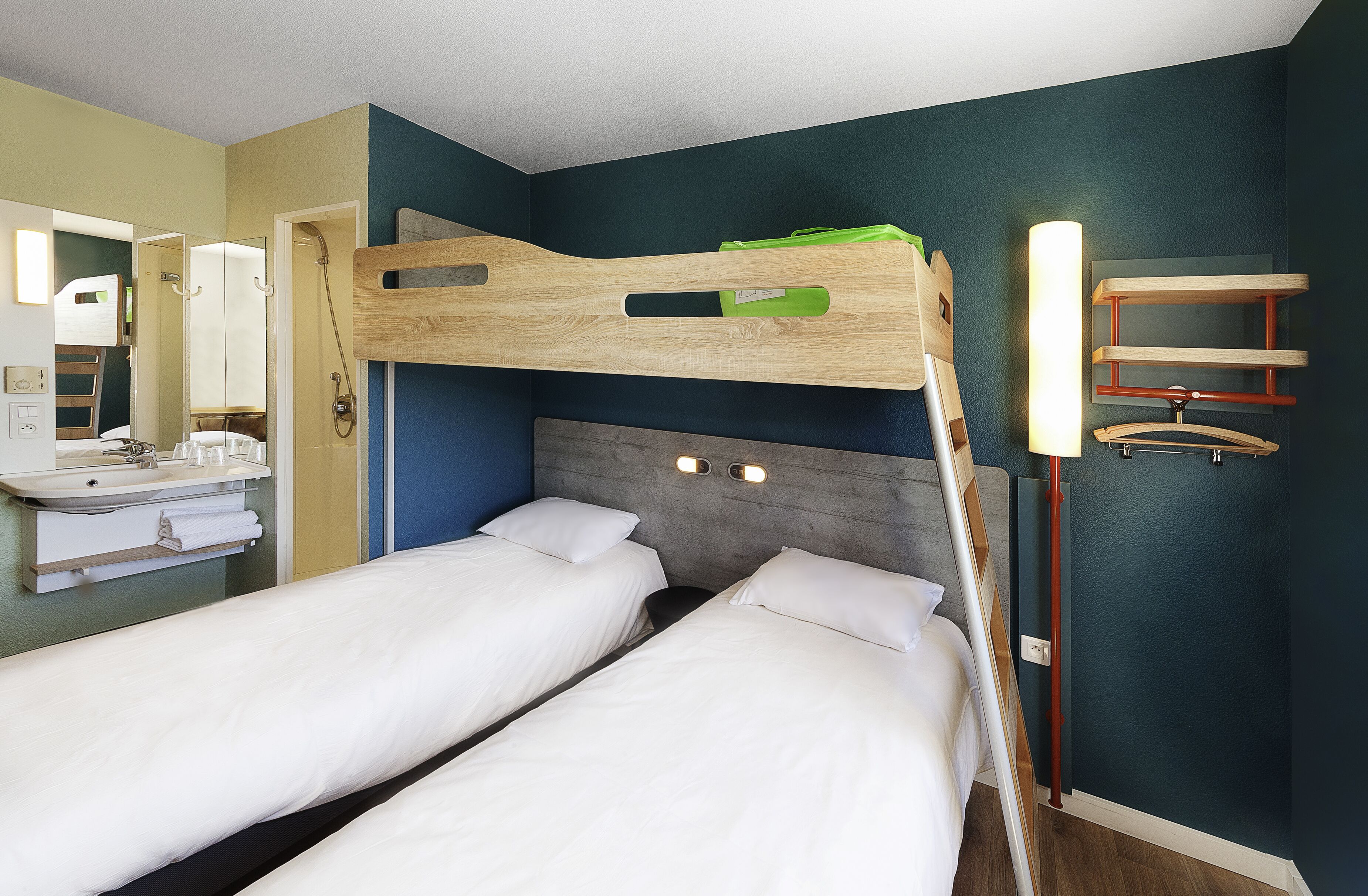 A hotel room with bunk beds, a bathroom with a sink and mirror, and a shelf with hanging items.