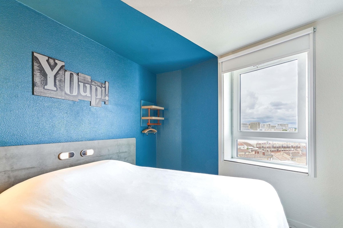 A bedroom with a bed, a blue accent wall, a window with a city view, and a wall-mounted shelf.