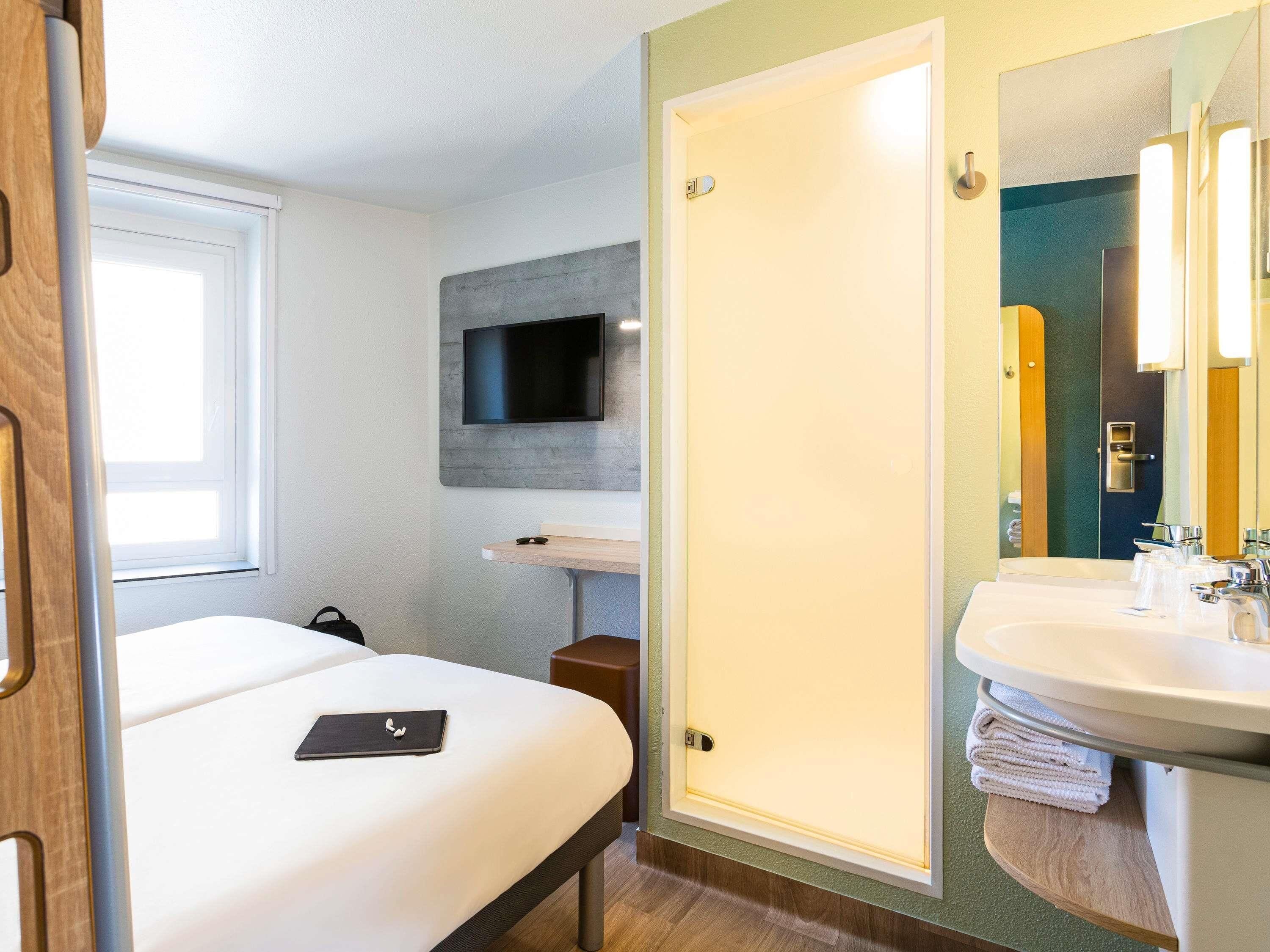 Habitación de hotel con cama, televisor montado en la pared, baño con lavabo y espejo, y puerta que da a otra habitación.