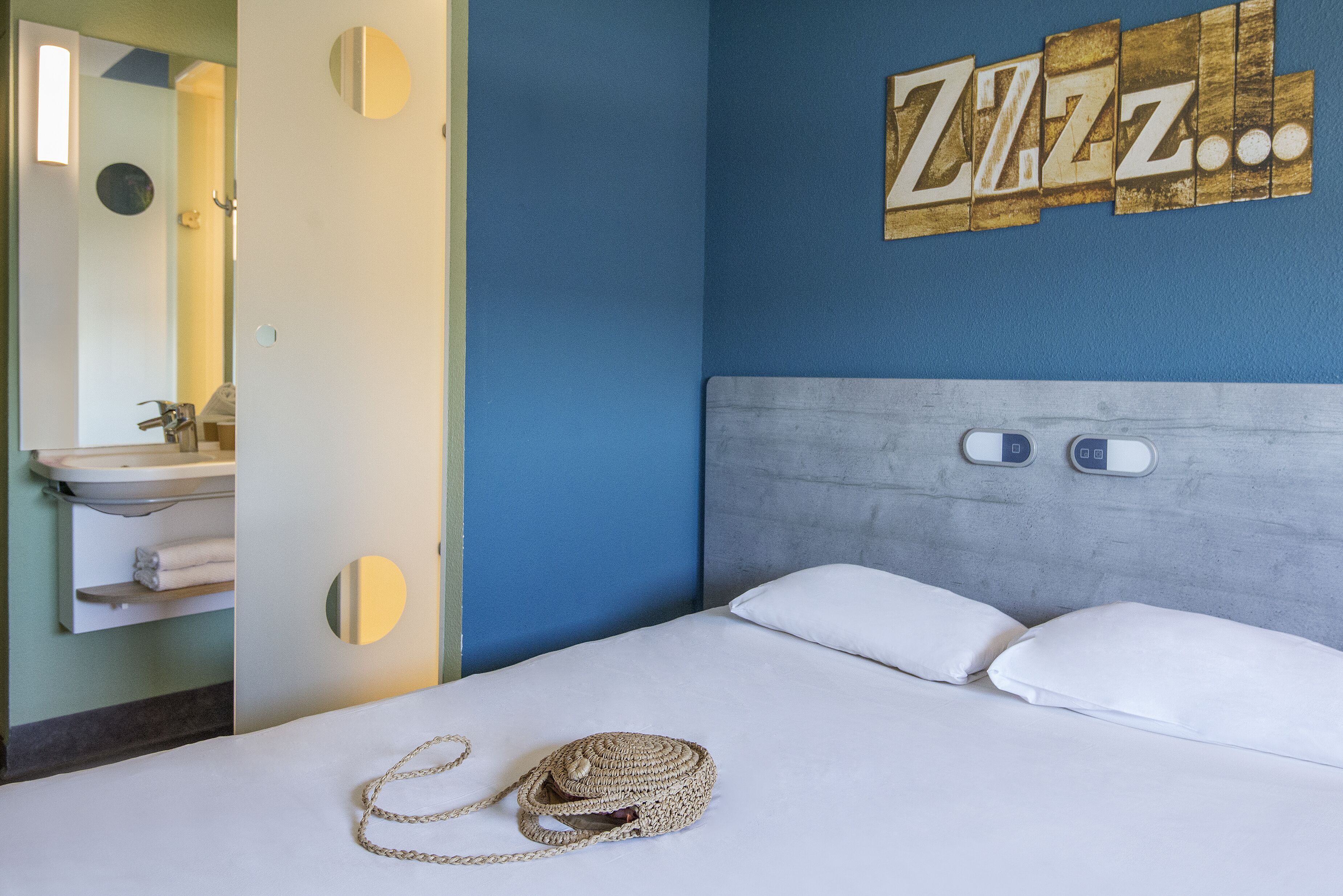 Une chambre d’hôtel avec un lit, une table de chevet, un miroir, une horloge murale et un panneau indiquant « Zzz… ».