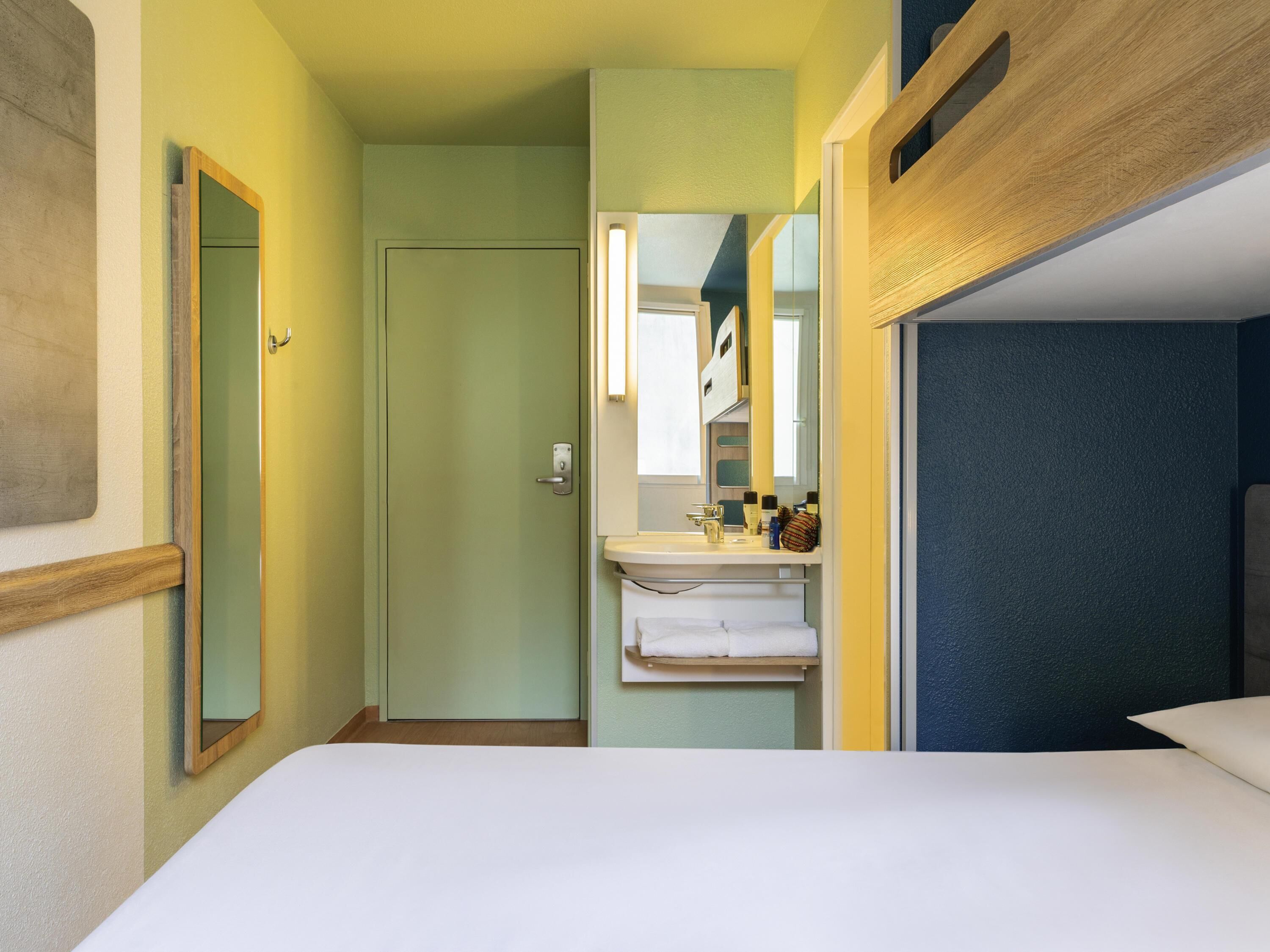 Habitación de hotel pequeña con una cama individual, baño con lavamanos y espejo, y una puerta que da a otra habitación.