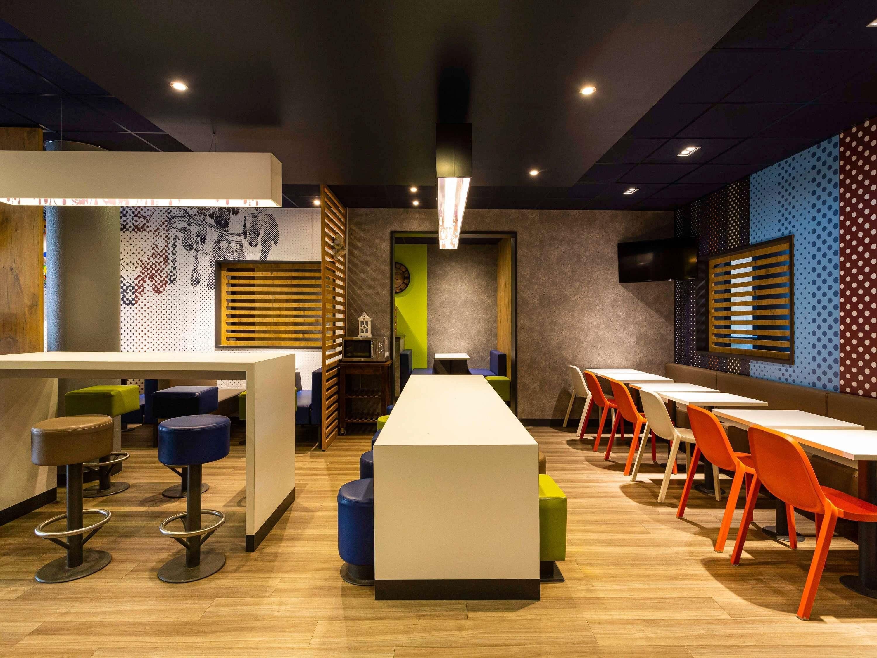 Un interior de restaurante moderno con mesas altas y sillas coloridas, un separador de madera y una pared con diseño.
