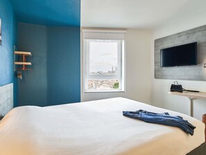 A hotel room with a bed, a window with a city view, a mounted TV, and a small shelf. - ibis budget Bordeaux Centre Gare Saint Jean (Bordeaux)